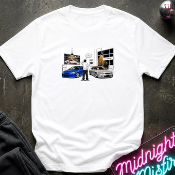 A white short-sleeve crewneck t-shirt laid flat on a dark textured surface. The center of the shirt features a graphic print depicting a person standing between a blue car and a silver car in front of a building.