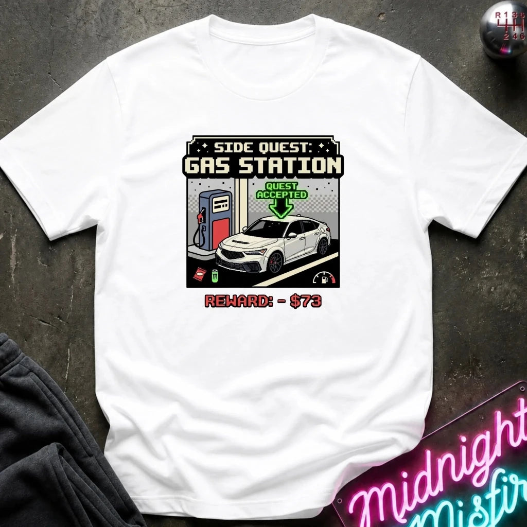 A white short-sleeve graphic t-shirt laid flat on a dark textured surface. The graphic depicts a retro video game side quest for a gas station with a white car, a green 'Quest Accepted' arrow, and red text reading 'Reward: $73'.