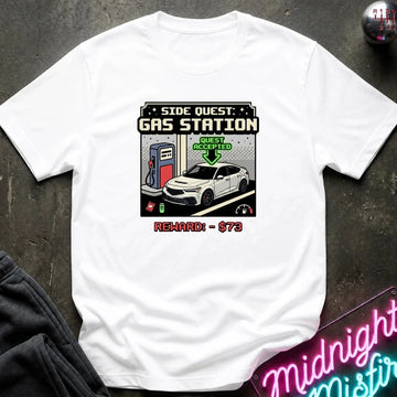 A white short-sleeve graphic t-shirt laid flat on a dark textured surface. The graphic depicts a retro video game side quest for a gas station with a white car, a green 'Quest Accepted' arrow, and red text reading 'Reward: $73'.