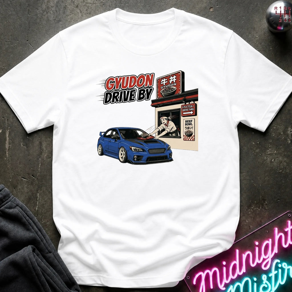 A white short-sleeve graphic t-shirt laid flat on a dark textured surface. The graphic features a blue sports car in front of a Japanese-style shop with the text 'GYUDON DRIVE BY'.