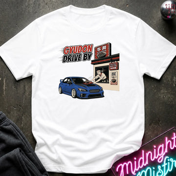 A white short-sleeve graphic t-shirt laid flat on a dark textured surface. The graphic features a blue sports car in front of a Japanese-style shop with the text 'GYUDON DRIVE BY'.