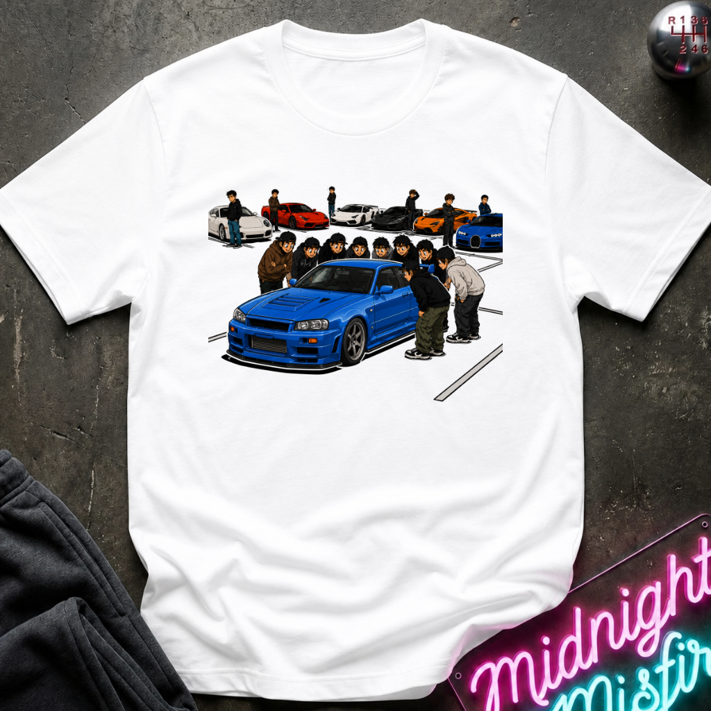 A white short-sleeve t-shirt laid flat on a dark textured surface, featuring a colorful graphic print of a blue sports car and a group of people in the center, with smaller cars in the background. A neon sign and dark clothing are partially visible around the edges.