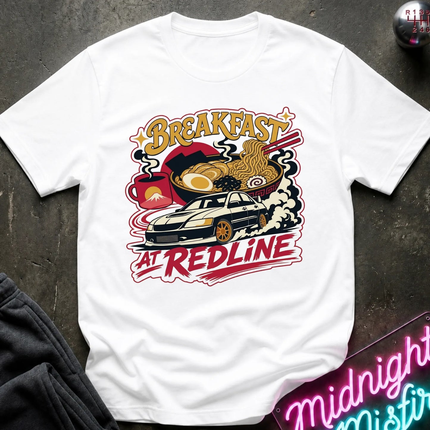A white short-sleeve graphic T-shirt laid flat on a dark textured surface. The shirt features a colorful retro-style print with the words “Breakfast at Redline,” a white car, ramen noodles, an egg, and red abstract shapes.