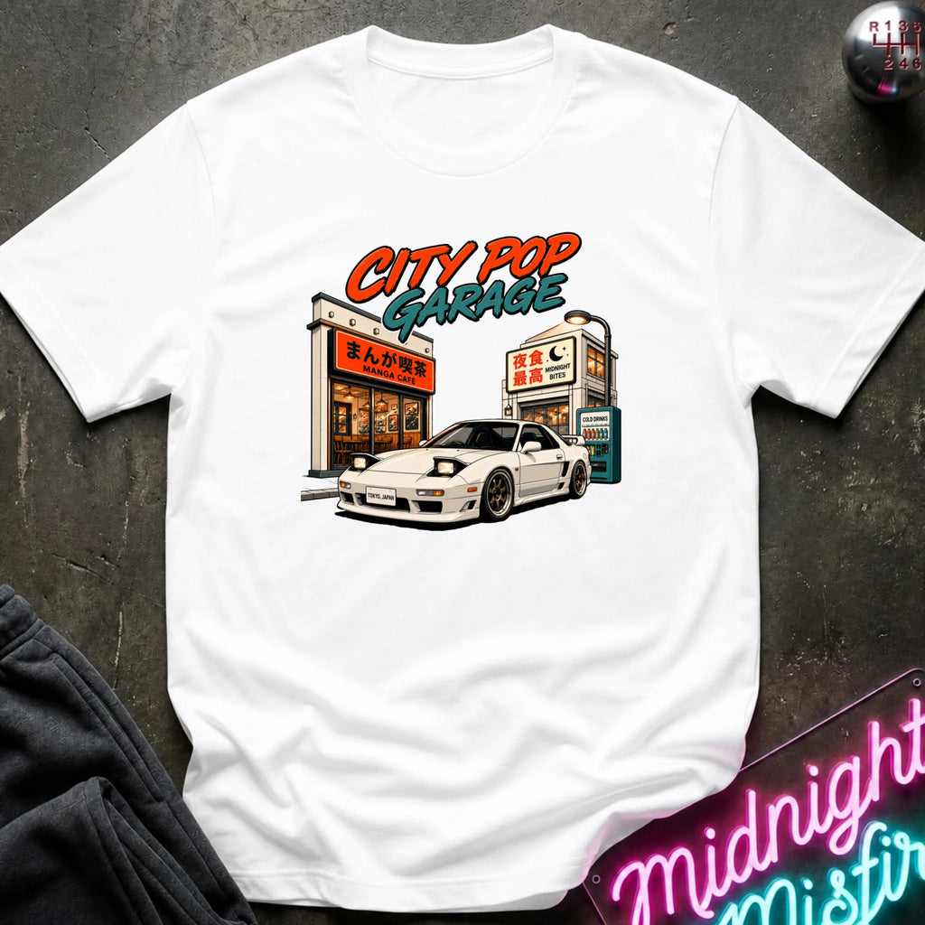 A white short-sleeve graphic t-shirt laid flat on a dark textured surface. The graphic features a white sports car parked in front of a cafe with the text 'CITY POP GARAGE' in orange and teal.