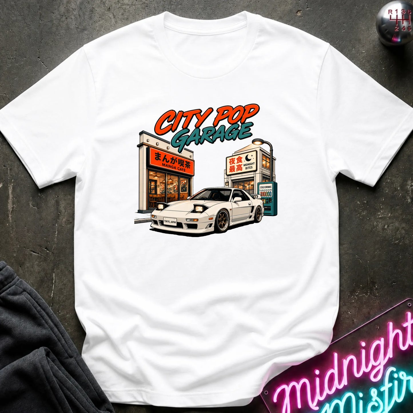 A white short-sleeve graphic t-shirt laid flat on a dark textured surface. The graphic features a white sports car parked in front of a cafe with the text 'CITY POP GARAGE' in orange and teal.