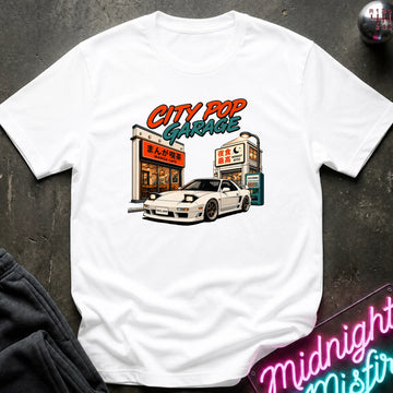 A white short-sleeve graphic t-shirt laid flat on a dark textured surface. The graphic features a white sports car parked in front of a cafe with the text 'CITY POP GARAGE' in orange and teal.