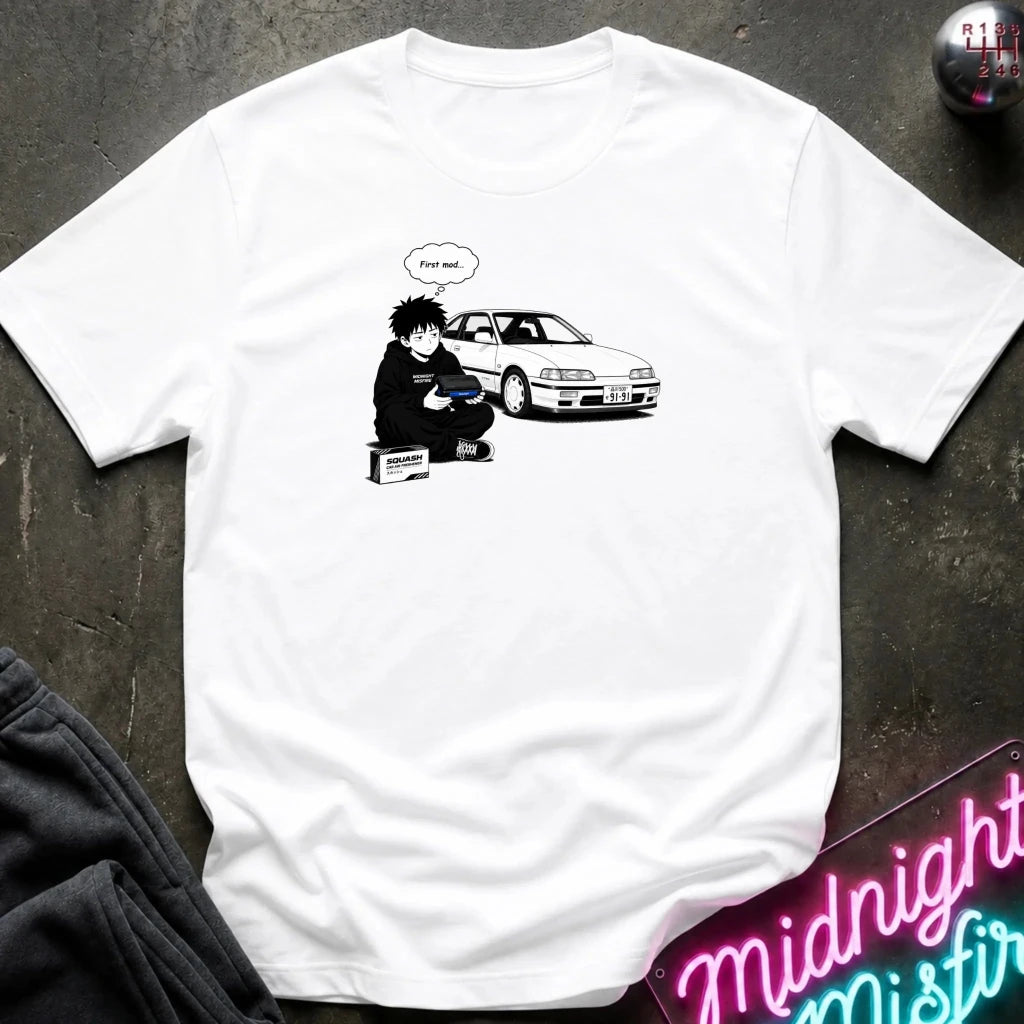 A white short-sleeve graphic t-shirt laid flat on a dark textured surface. The graphic shows a black-and-white illustration of a person sitting next to a white car with a speech bubble