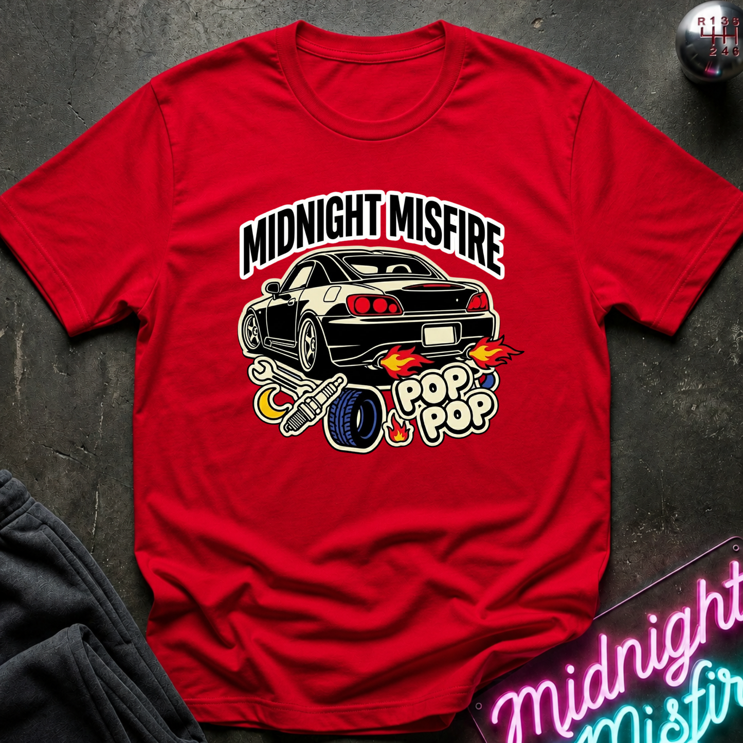 A red short-sleeve graphic T-shirt laid flat on a dark textured surface. The shirt features a large front print with the words “MIDNIGHT MISFIRE” above a black sports car, flames, a tire, and “POP POP” text.