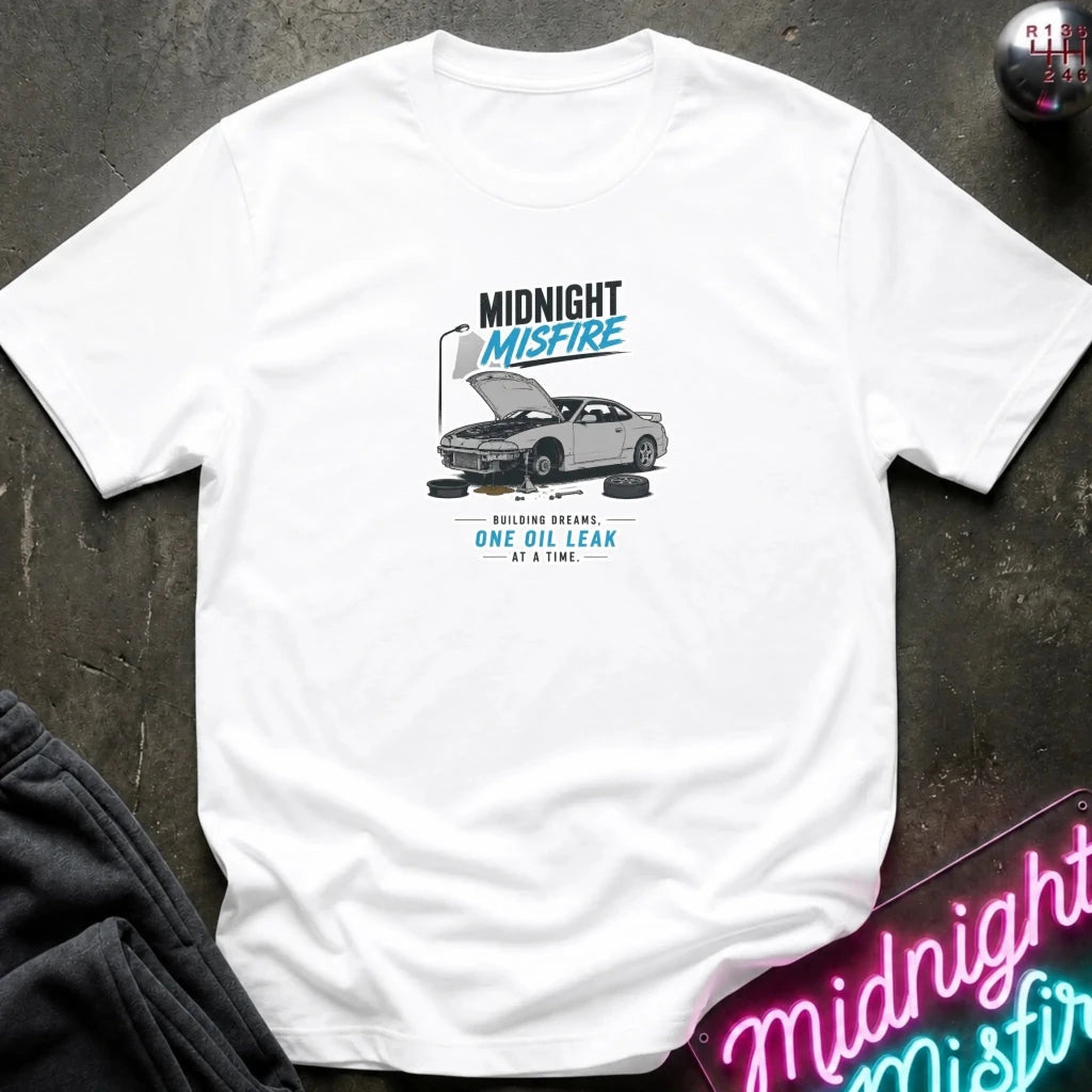 A white short-sleeve graphic T-shirt laid flat on a dark textured surface. The shirt features a printed design of a gray car with its hood open, accompanied by the text 'MIDNIGHT MISFIRE' and 'BUILDING BREAMS ONE OIL LEAK AT A TIME.