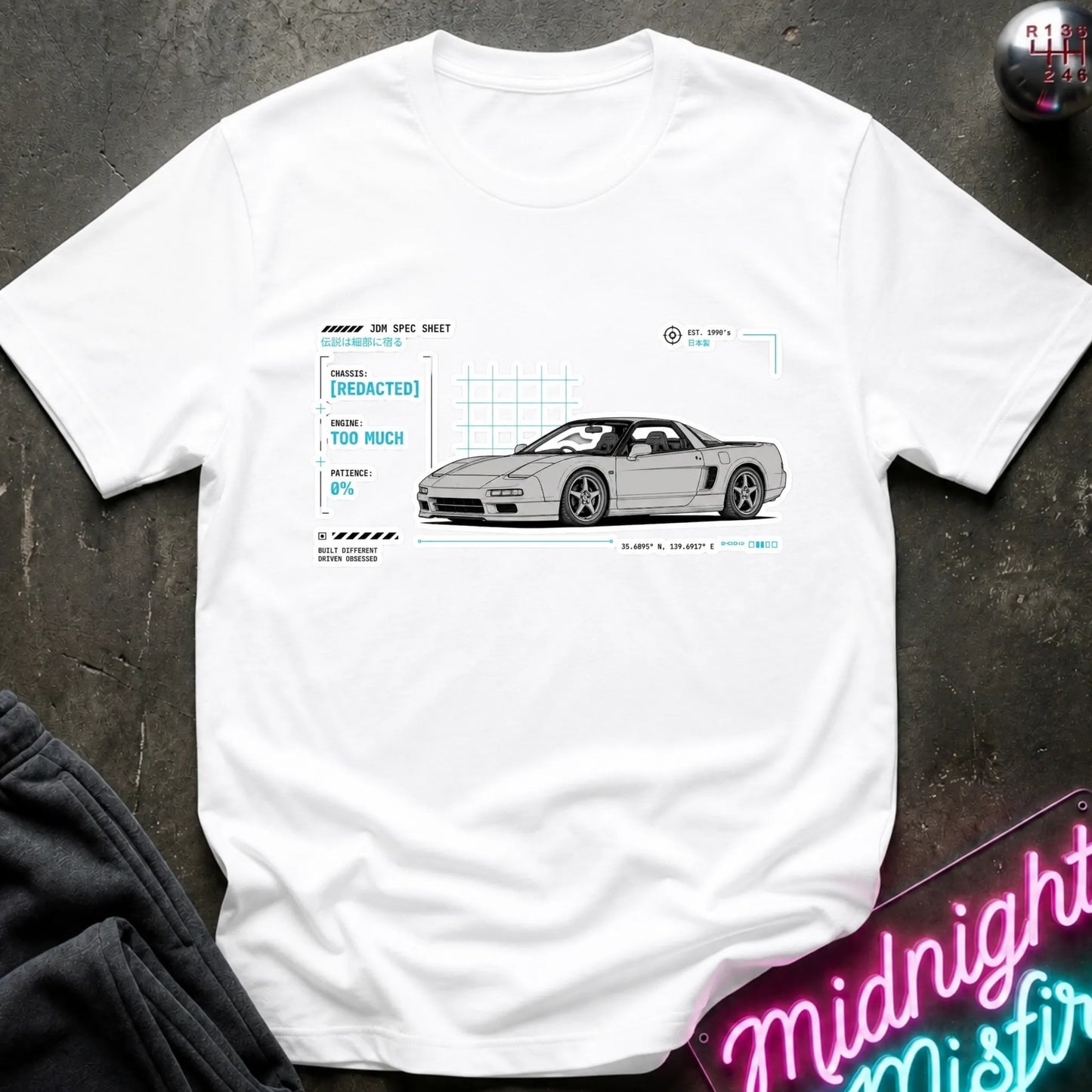 A white short-sleeve t-shirt laid flat on a dark textured surface, featuring a white and light blue graphic print of a sports car with technical-style labels. A neon sign and folded dark fabric are partially visible around the edges.