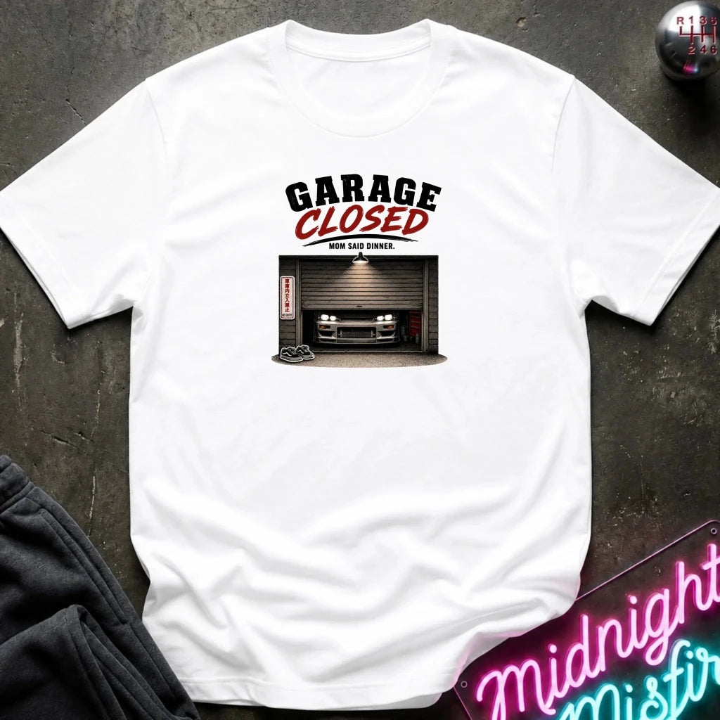 A white short-sleeve graphic t-shirt laid flat on a dark textured surface. The graphic features the text 'GARAGE CLOSED' in black and red, above an illustration of a car parked in front of a closed garage door.