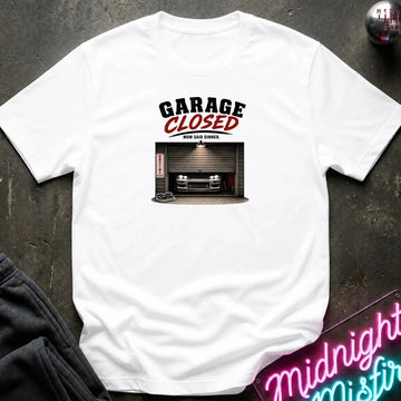 A white short-sleeve graphic t-shirt laid flat on a dark textured surface. The graphic features the text 'GARAGE CLOSED' in black and red, above an illustration of a car parked in front of a closed garage door.