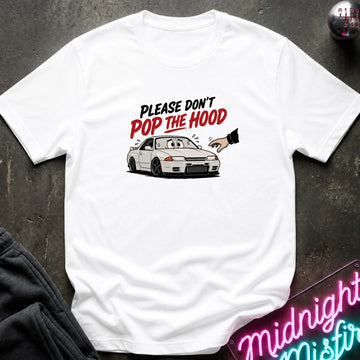A white short-sleeve graphic t-shirt laid flat on a dark textured surface. The graphic features a white sports car with the text 'PLEASE DON'T POP THE HOOD' in black and red lettering, with a cartoon hand pointing toward the hood.