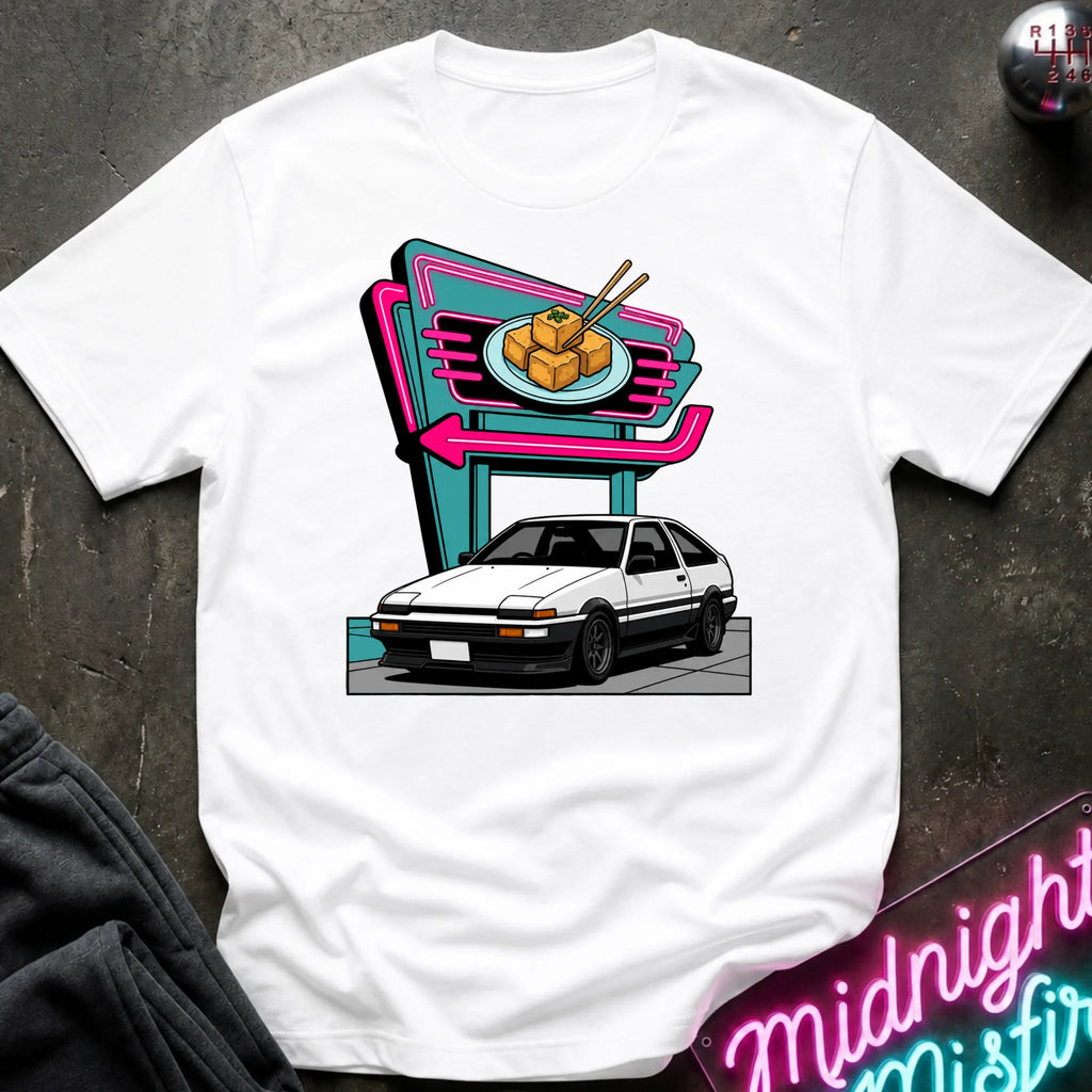 A white short-sleeve crewneck t-shirt featuring a retro-style graphic of a white sports car parked in front of a neon sign. The shirt is displayed flat on a dark textured surface with a dark sweatshirt and a neon sign partially visible as props.