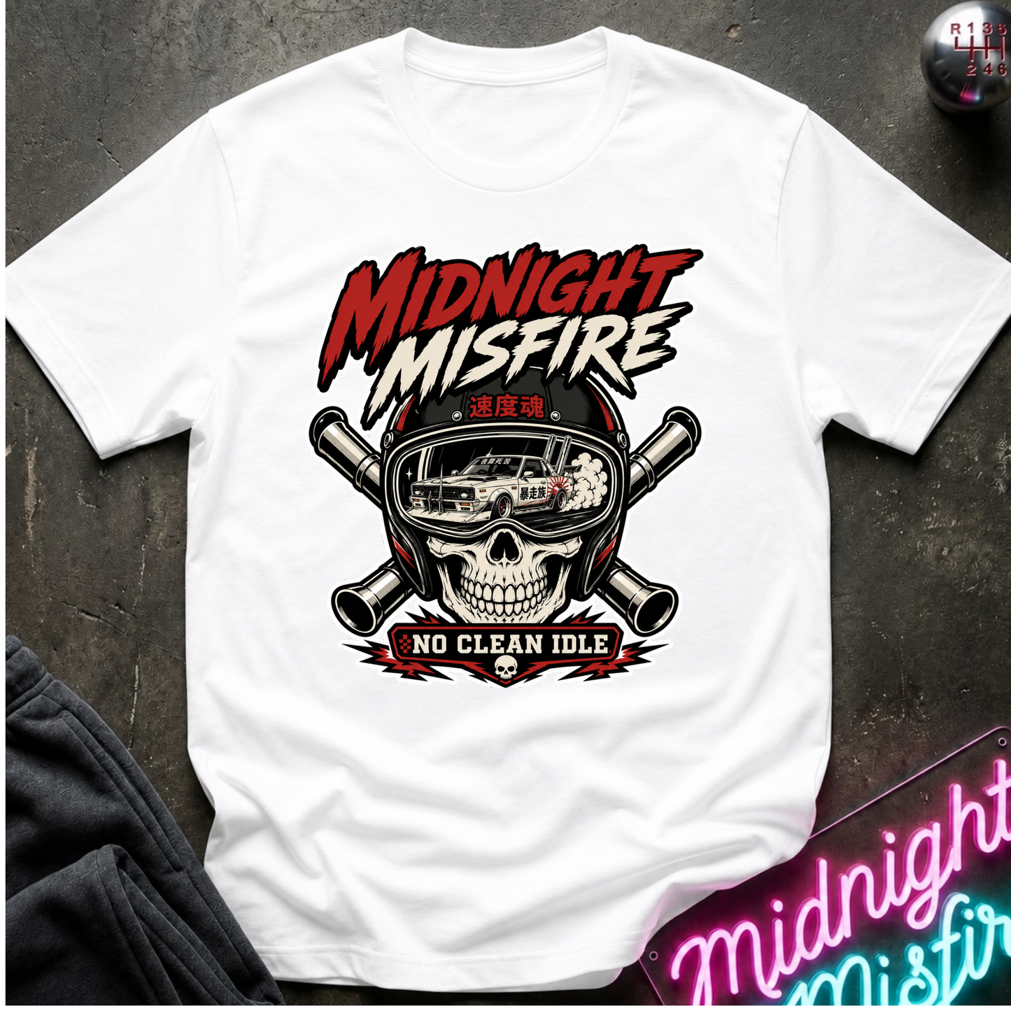 A white short-sleeve graphic T-shirt laid flat on a dark textured surface. The shirt features a large front print with the words “MIDNIGHT MISFIRE” in red and white, a skull wearing a helmet with crossed tools behind it, and “NO CLEAN IDLE” below.