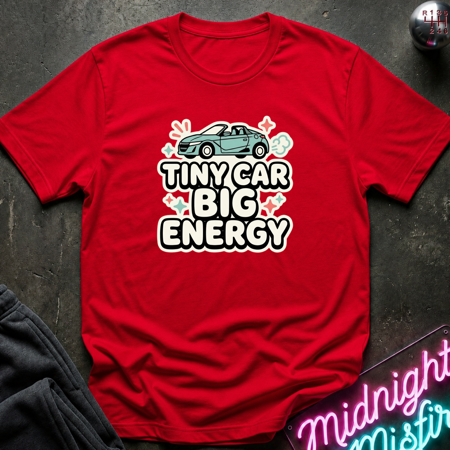 A red short-sleeve graphic T-shirt laid flat on a dark textured surface. The shirt features a light blue convertible car illustration above the bold white text 'TINY CAR BIG ENERGY' with small pink and white decorative accents.