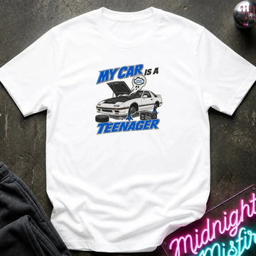 A white short-sleeve graphic t-shirt laid flat on a dark textured surface. The graphic features a white sports car with its hood open, blue text reading 'MY CAR IS A TEENAGER'
