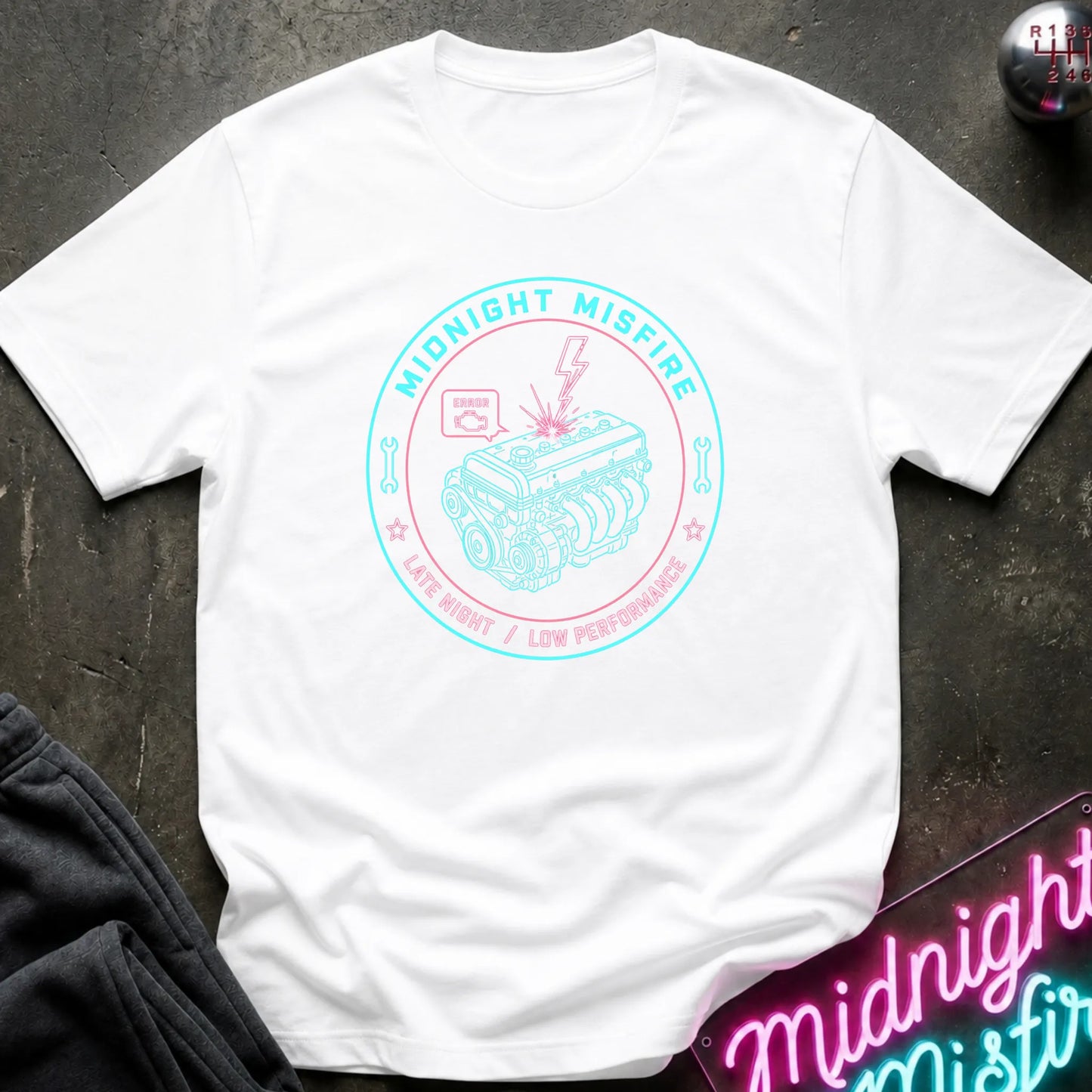 A white short-sleeve t-shirt with a circular graphic on the chest reading 'Midnight Misfire' and showing a pink and teal engine illustration, laid flat on a dark textured surface with a neon sign and dark pants partially visible.