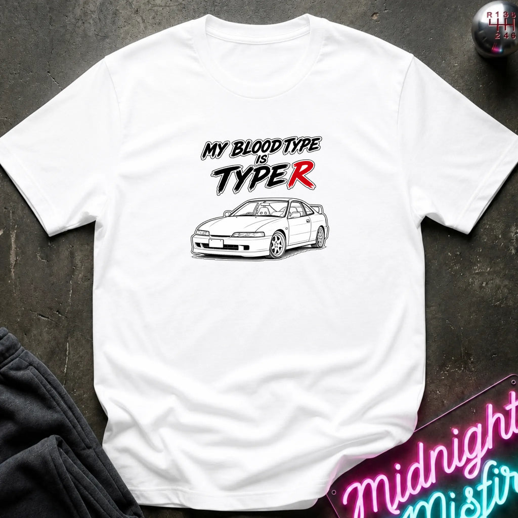 A white short-sleeve graphic T-shirt laid flat on a dark textured surface. The shirt features the text 'MY BLOOD TYPE IS TYPE R' in black and red, above a black-and-white line drawing of a sports car.