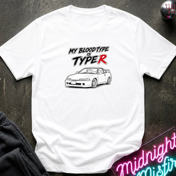 A white short-sleeve graphic T-shirt laid flat on a dark textured surface. The shirt features the text 'MY BLOOD TYPE IS TYPE R' in black and red, above a black-and-white line drawing of a sports car.