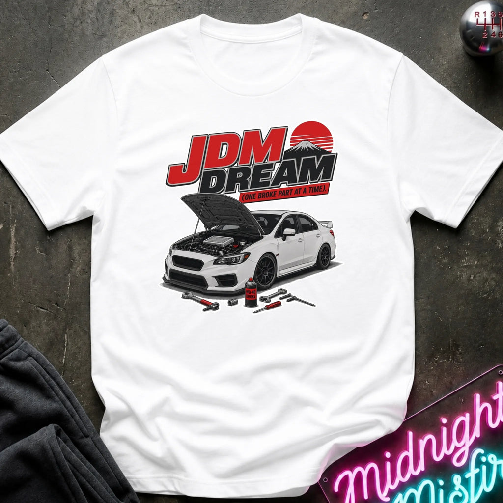A white short-sleeve graphic T-shirt laid flat on a dark textured surface. The shirt features a large front print with the text 'JDM DREAM' in red and black above an illustration of a white sports car with its hood open, plus small automotive tools.