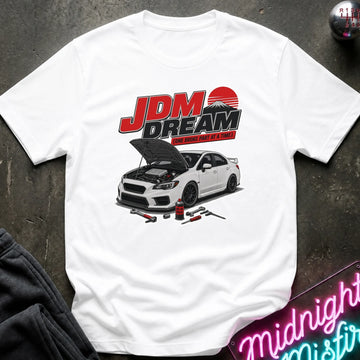 A white short-sleeve graphic T-shirt laid flat on a dark textured surface. The shirt features a large front print with the text 'JDM DREAM' in red and black above an illustration of a white sports car with its hood open, plus small automotive tools.