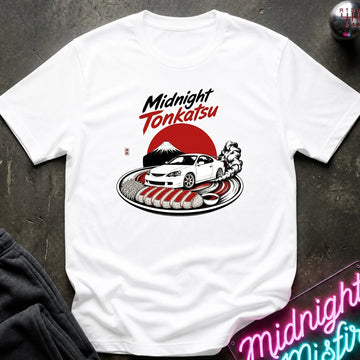 A white short-sleeve graphic T-shirt laid flat on a dark textured surface. The shirt features a stylized graphic with the words 'Midnight Tonkatsu' in white and red, a white sports car, a red sun, a black-and-white figure, and a plate of fried cutlets.