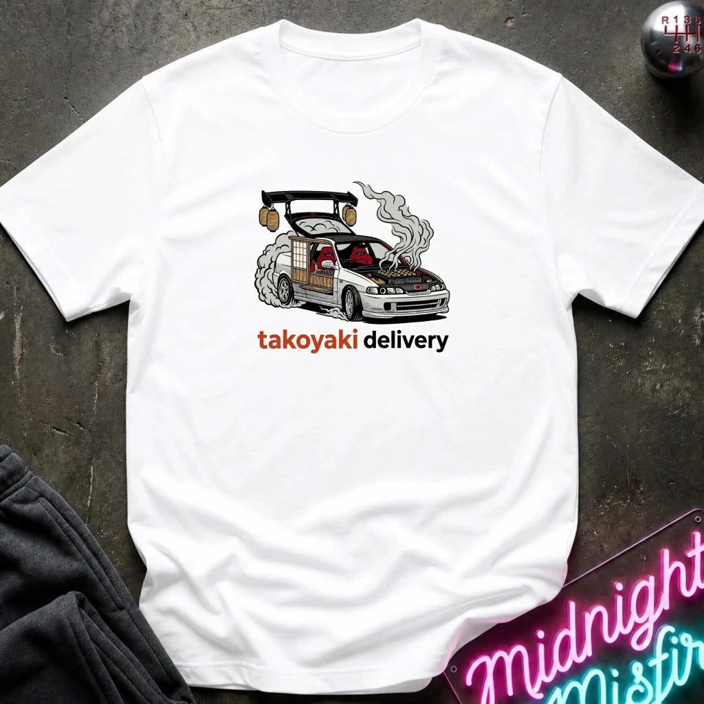 A white short-sleeve t-shirt laid flat on a dark textured surface, featuring a graphic of a smoking white sports car with the text 'takoyaki delivery' in orange and black. A neon sign and dark clothing are partially visible in the corners as props.