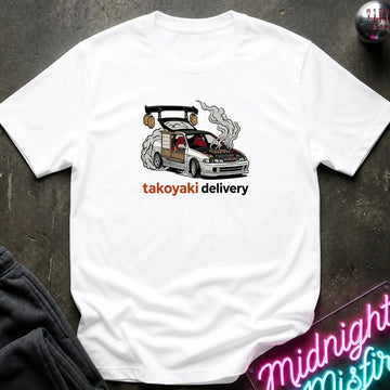 A white short-sleeve t-shirt laid flat on a dark textured surface, featuring a graphic of a smoking white sports car with the text 'takoyaki delivery' in orange and black. A neon sign and dark clothing are partially visible in the corners as props.
