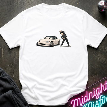 A white short-sleeve t-shirt laid flat on a dark textured surface, featuring a graphic print of a white sports car and a person in black standing next to it. A neon sign and folded dark clothing are partially visible as props.