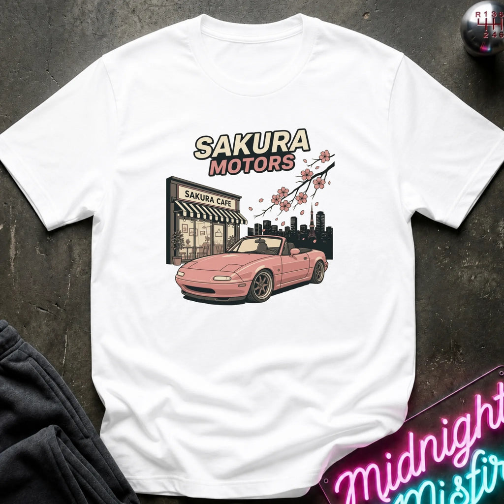 A white short-sleeve graphic T-shirt laid flat on a dark textured surface. The shirt features a printed design of a pink convertible car in front of a 'Sakura Motors' shop, with cherry blossoms and a city skyline.