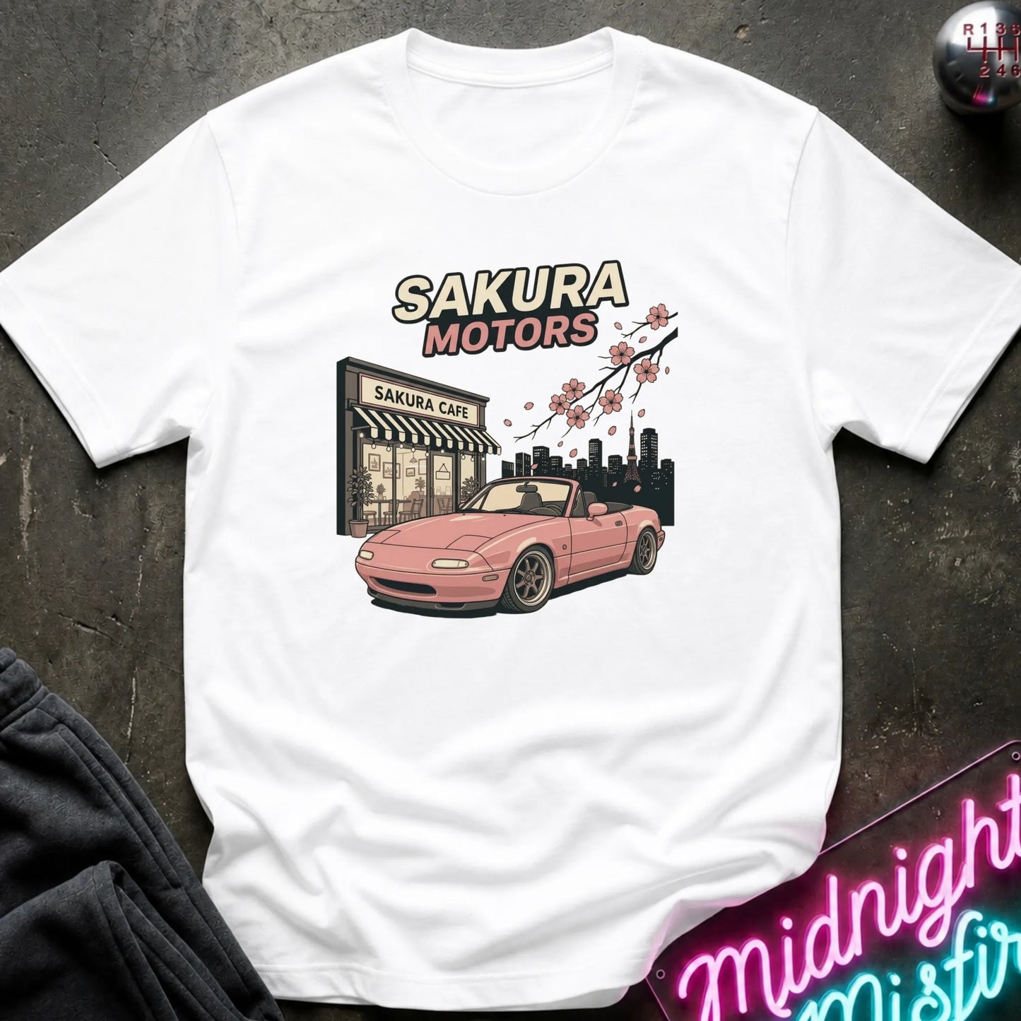 A white short-sleeve graphic T-shirt laid flat on a dark textured surface. The shirt features a printed design of a pink convertible car in front of a 'Sakura Motors' shop, with cherry blossoms and a city skyline.