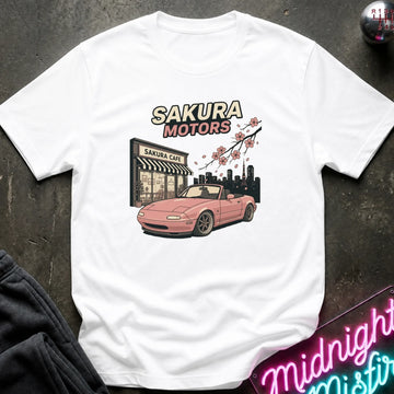 A white short-sleeve graphic T-shirt laid flat on a dark textured surface. The shirt features a printed design of a pink convertible car in front of a 'Sakura Motors' shop, with cherry blossoms and a city skyline.