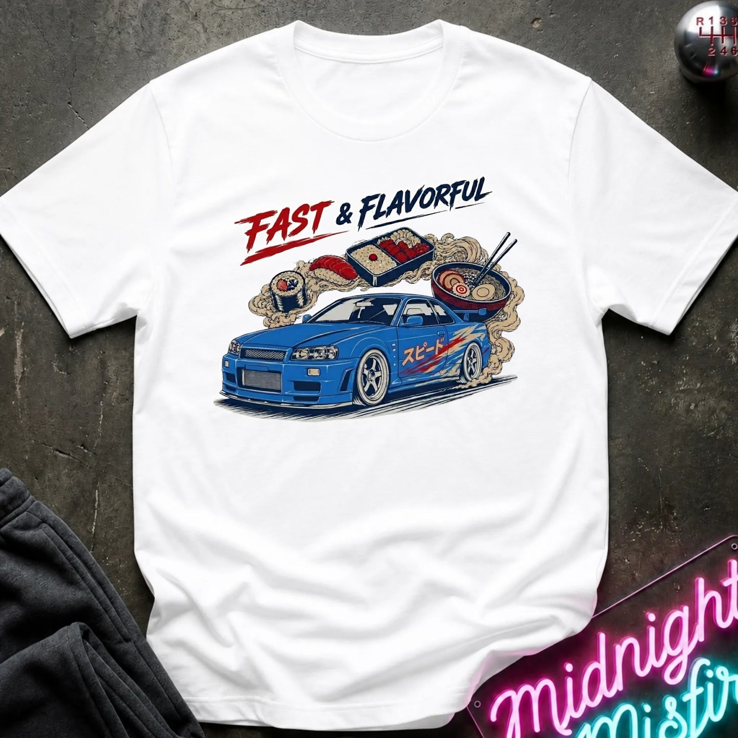 A white short-sleeve graphic T-shirt laid flat on a dark textured surface. The shirt features a colorful print of a blue sports car with ramen noodles and chopsticks, plus the text 'FAST & FLAVORFUL' in red and black.