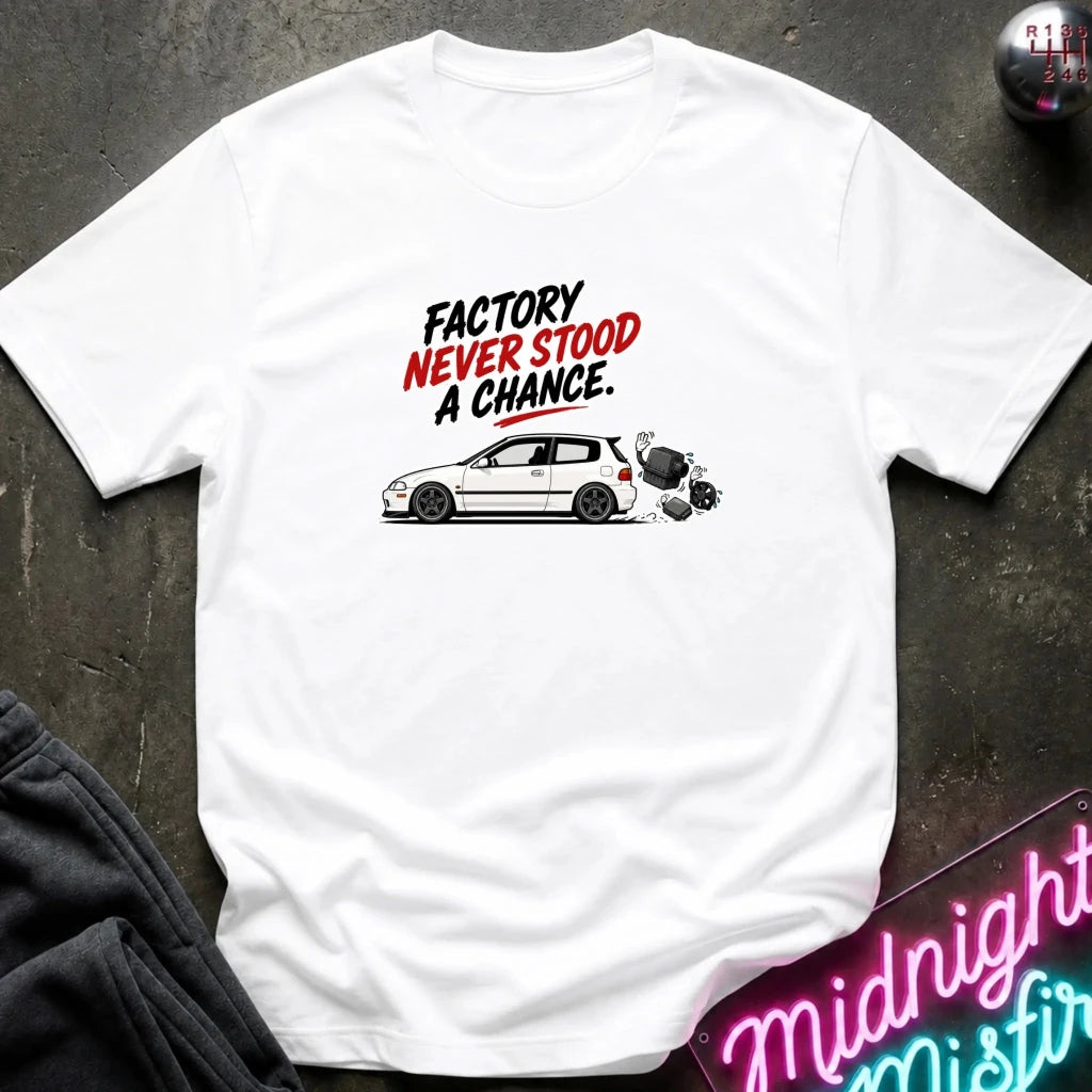A white short-sleeve graphic T-shirt laid flat on a dark textured surface. The shirt features a printed design with the text 'FACTORY NEVER STOOD A CHANCE.
