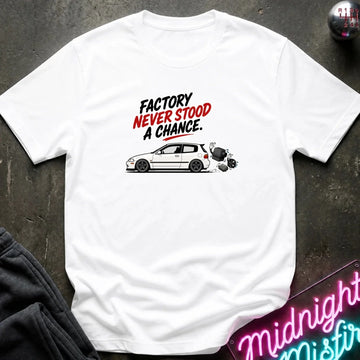 A white short-sleeve graphic T-shirt laid flat on a dark textured surface. The shirt features a printed design with the text 'FACTORY NEVER STOOD A CHANCE.