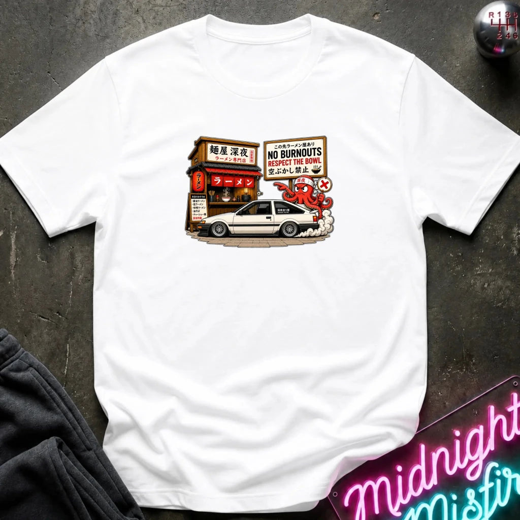A white short-sleeve t-shirt laid flat on a dark textured surface, featuring a graphic print of a white sports car parked in front of a Japanese street shop with signs reading 'NO BURNOUTS'.