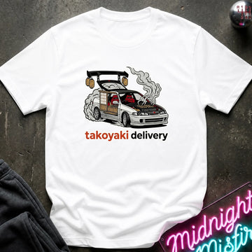 A white short-sleeve graphic T-shirt laid flat on a dark textured surface. The shirt features a cartoon illustration of a smoking car with the text 'takoyaki delivery' printed below it in orange and black.