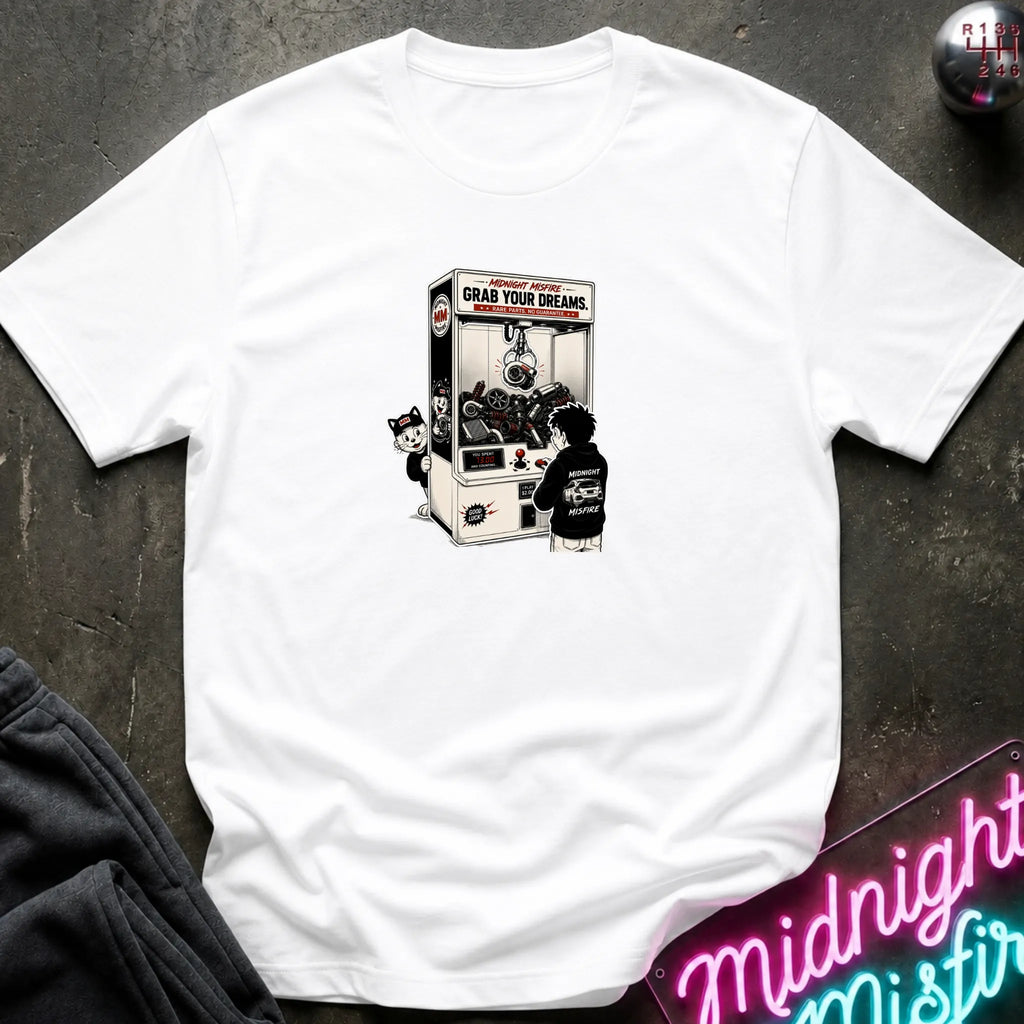 A white short-sleeve crewneck t-shirt laid flat on a dark textured surface, featuring a central graphic of a person in a black hoodie playing a claw machine. 