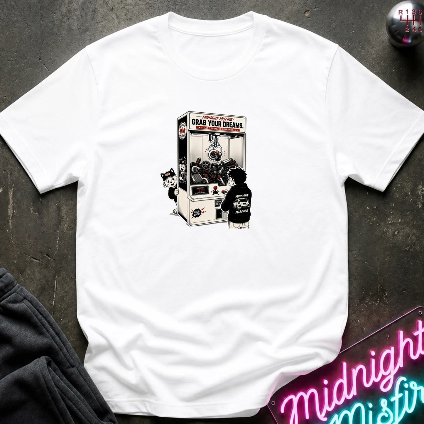 A white short-sleeve crewneck t-shirt laid flat on a dark textured surface, featuring a central graphic of a person in a black hoodie playing a claw machine. 