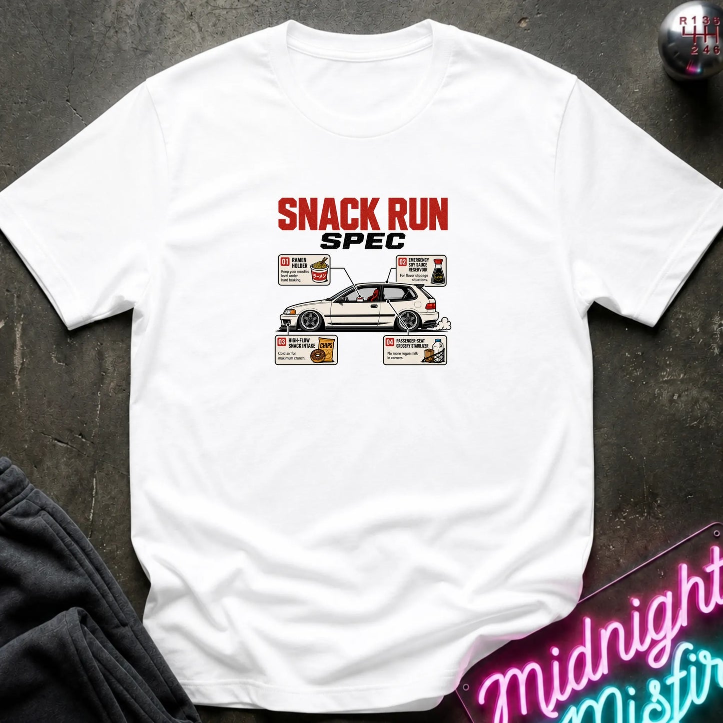 A white short-sleeve graphic T-shirt laid flat on a dark textured surface. The shirt features a red and black 'SNACK RUN SPEC' text graphic above a white race car with detailed nutrition labels around it.