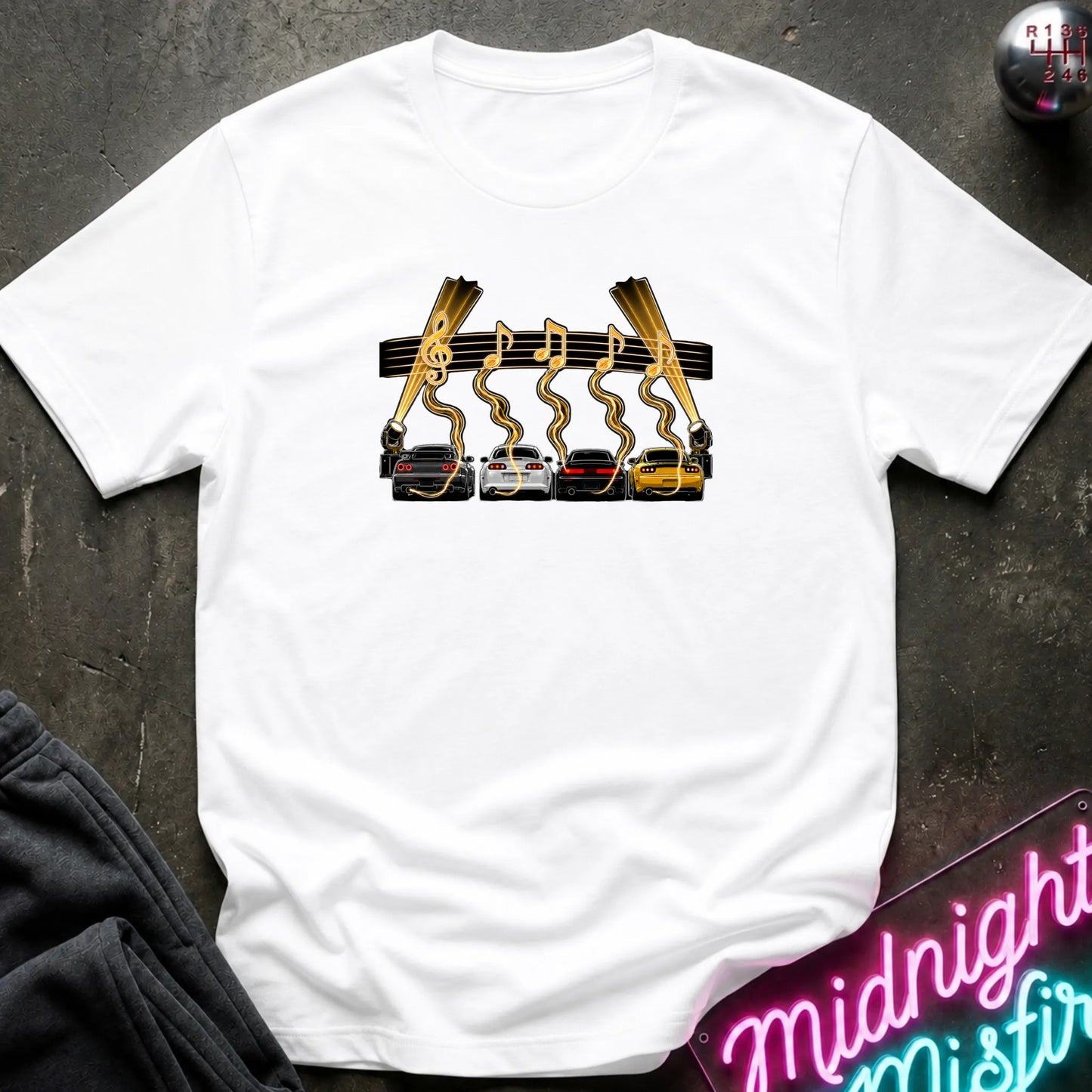 A white short-sleeve t-shirt laid flat on a dark textured surface, featuring a graphic print of four cars (red, white, gray, yellow) with gold wavy lines and musical notes above them, flanked by two  lights.