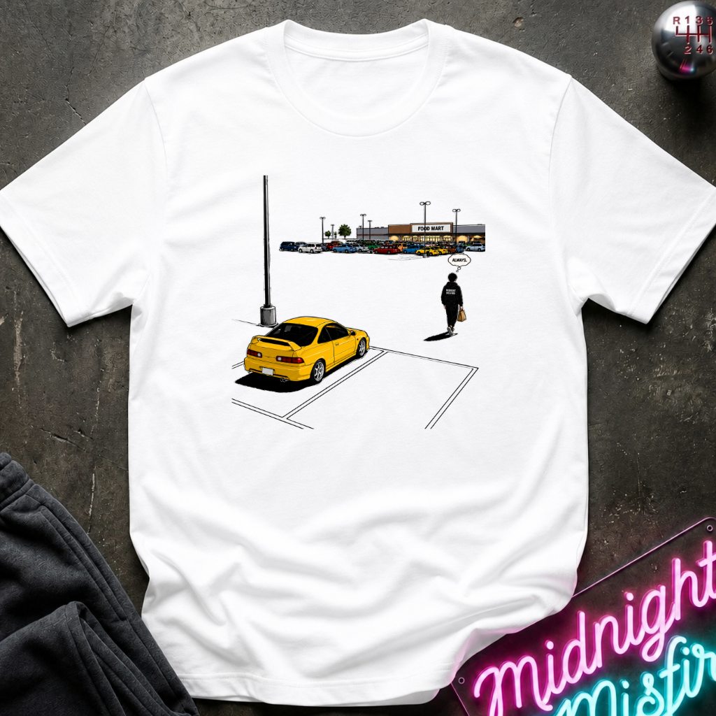 A white short-sleeve graphic t-shirt laid flat on a dark textured surface. The graphic shows a yellow car, a pedestrian, and a parking structure.