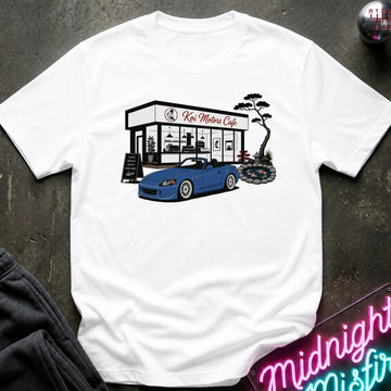 A white short-sleeve t-shirt laid flat on a dark textured surface, featuring a graphic print of a blue convertible sports car parked in front of 'Koi Motor Cafe' with a black tree illustration.