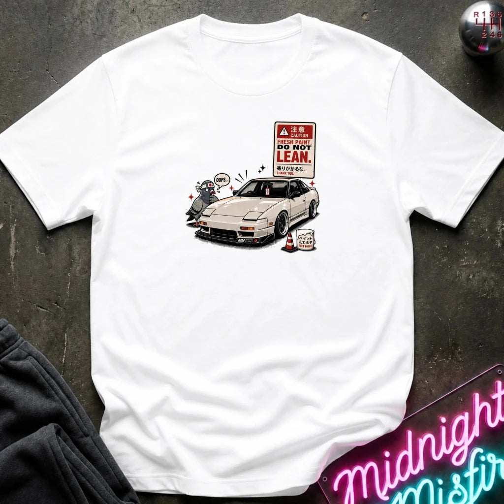 A white short-sleeve t-shirt laid flat on a dark textured surface, featuring a graphic print of a white sports car next to a 'DO NOT LEAN' warning sign.
