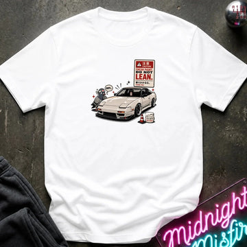 A white short-sleeve t-shirt laid flat on a dark textured surface, featuring a graphic print of a white sports car next to a 'DO NOT LEAN' warning sign.