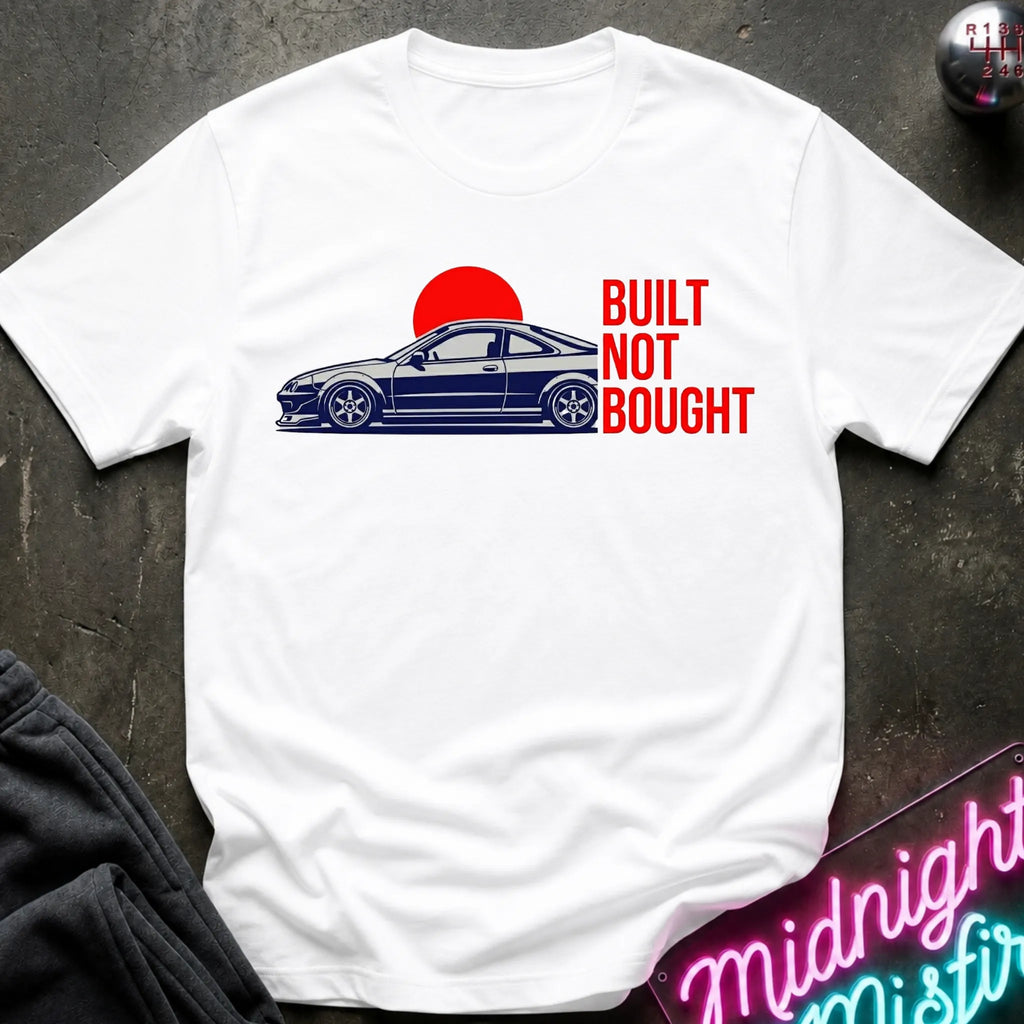 A white short-sleeve graphic T-shirt laid flat on a dark textured surface. The shirt features a side-profile illustration of a dark blue car in front of a red sun, with the text 'BUILT NOT BOUGHT' printed in red on the right side.