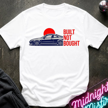 A white short-sleeve graphic T-shirt laid flat on a dark textured surface. The shirt features a side-profile illustration of a dark blue car in front of a red sun, with the text 'BUILT NOT BOUGHT' printed in red on the right side.