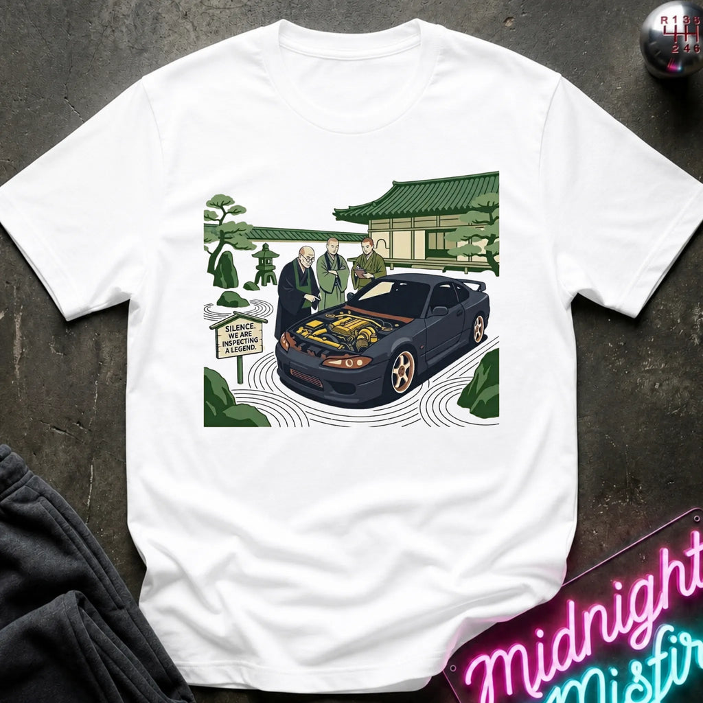 White short-sleeve graphic t-shirt laid flat on a dark textured surface. The graphic shows a black sports car with its hood open, surrounded by two men in suits, a Japanese-style building, and green trees.