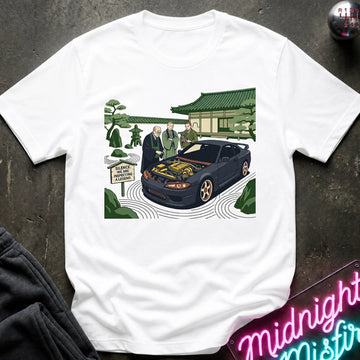 White short-sleeve graphic t-shirt laid flat on a dark textured surface. The graphic shows a black sports car with its hood open, surrounded by two men in suits, a Japanese-style building, and green trees.